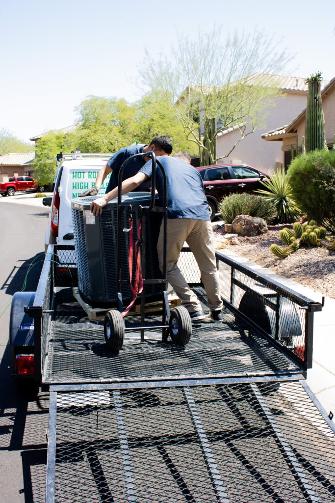 Legendary Energy technicians loading an air conditioning condenser onto a trailer behind the company van.