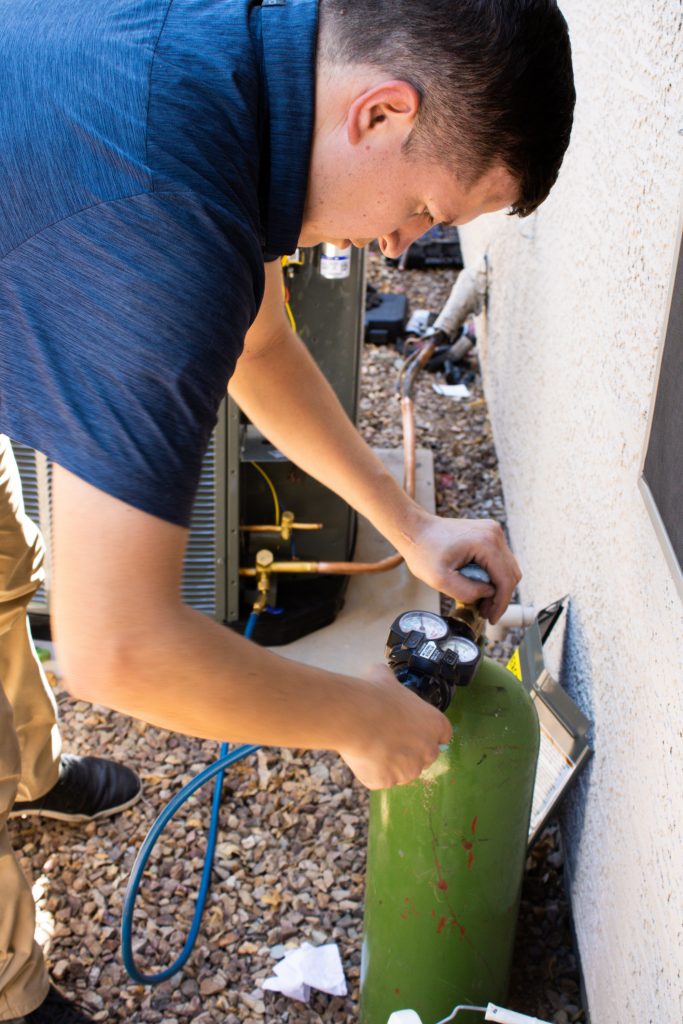 Legendary Energy technician connecting gauges to a refrigerant tank beside an outdoor unit