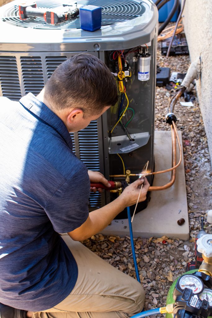 HVAC technician in a Legendary Energy shirt brazing copper refrigerant lines on an open air conditioning condenser